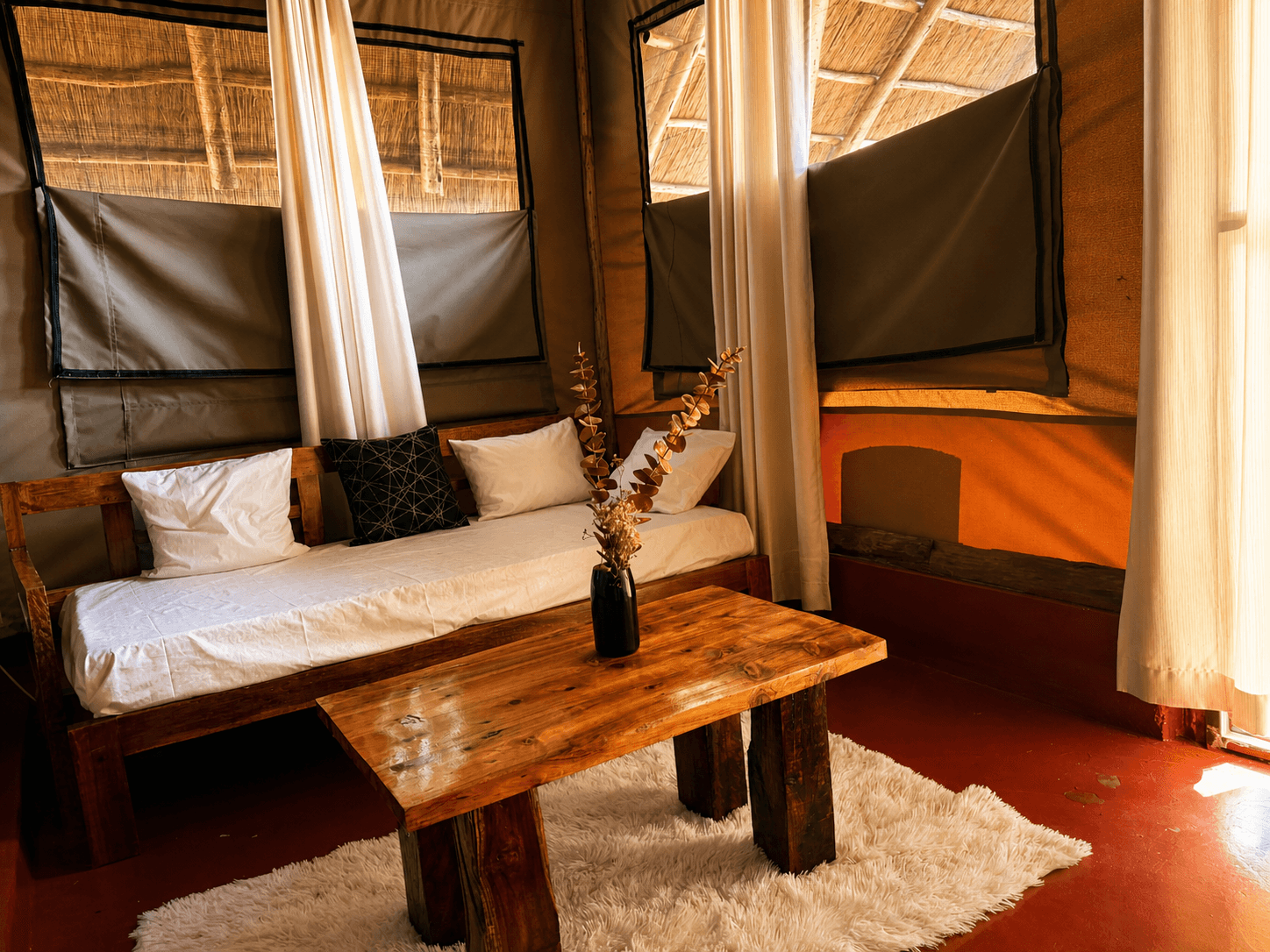 Luxury Safari Tent sitting area at The Elephant Oasis — wooden daybed with white cushions, live-edge coffee table, dried eucalyptus, canvas tent windows with sheer curtains, thatched ceiling, Murchison Falls Uganda