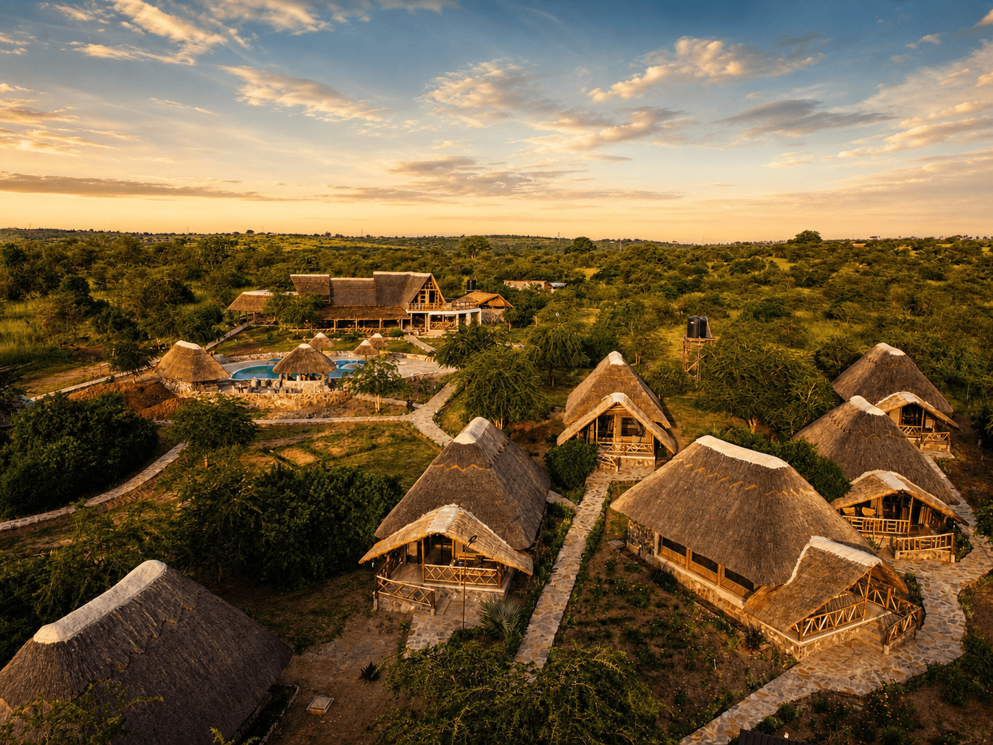Aerial view of The Elephant Oasis — multiple safari tent rooftops, elephant-shaped pool, stone paths, main lodge building, and open Ugandan savanna stretching to the horizon at golden hour