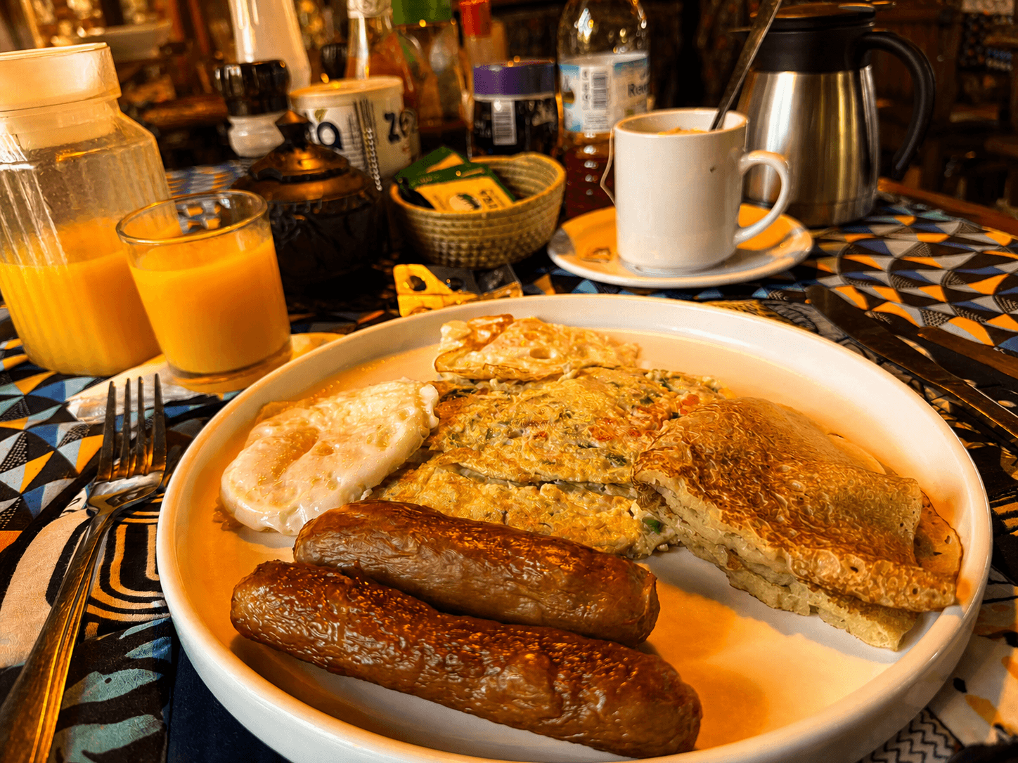 Real breakfast at The Elephant Oasis — plated omelette, fried egg, chicken sausages, orange juice, and coffee on an African-print tablecloth, Murchison Falls Uganda