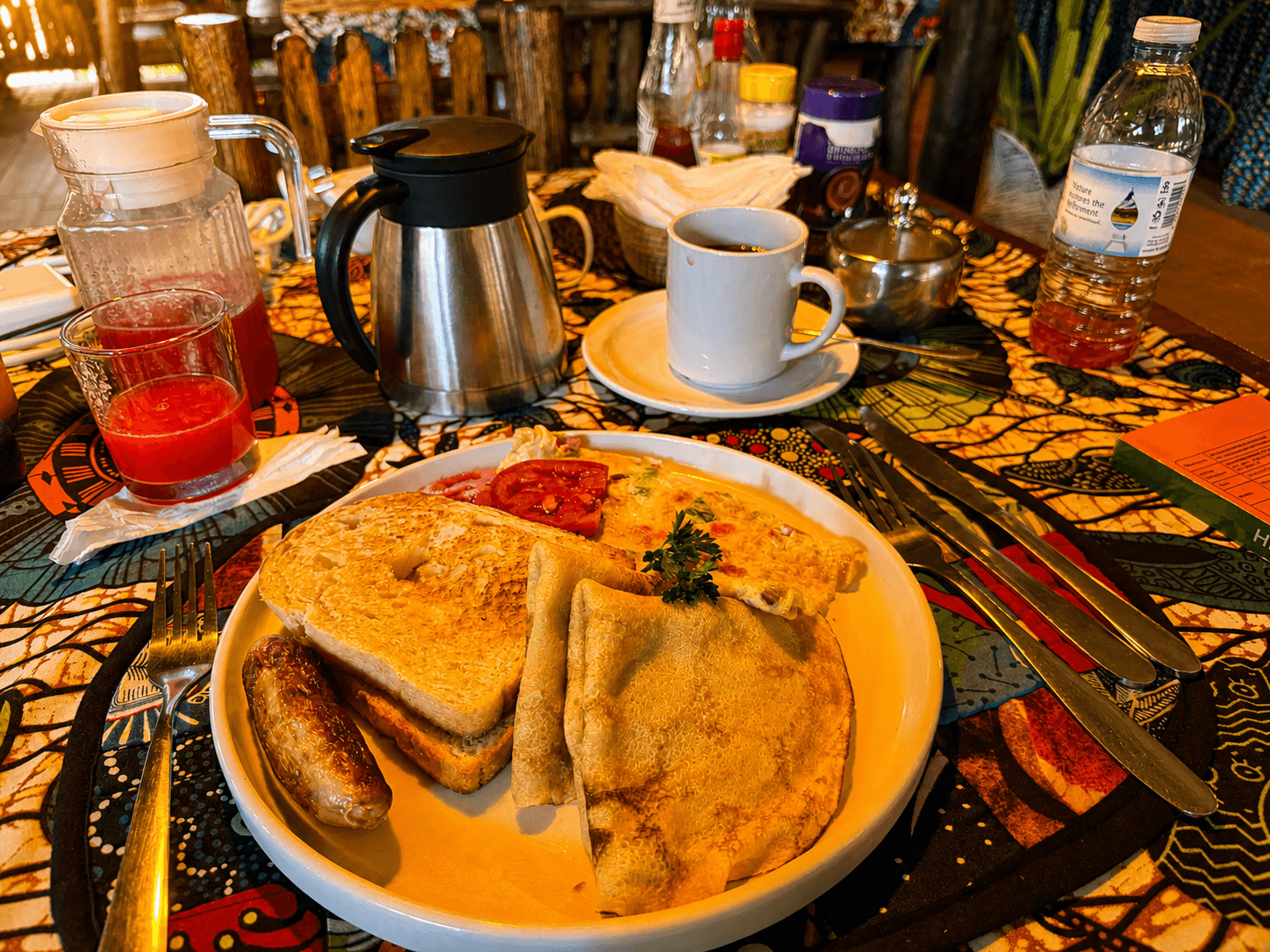 Breakfast table at The Elephant Oasis — pancake, sausage, toast, tomato, scrambled egg, juice, and coffee thermos on a patterned tablecloth, Murchison Falls Uganda