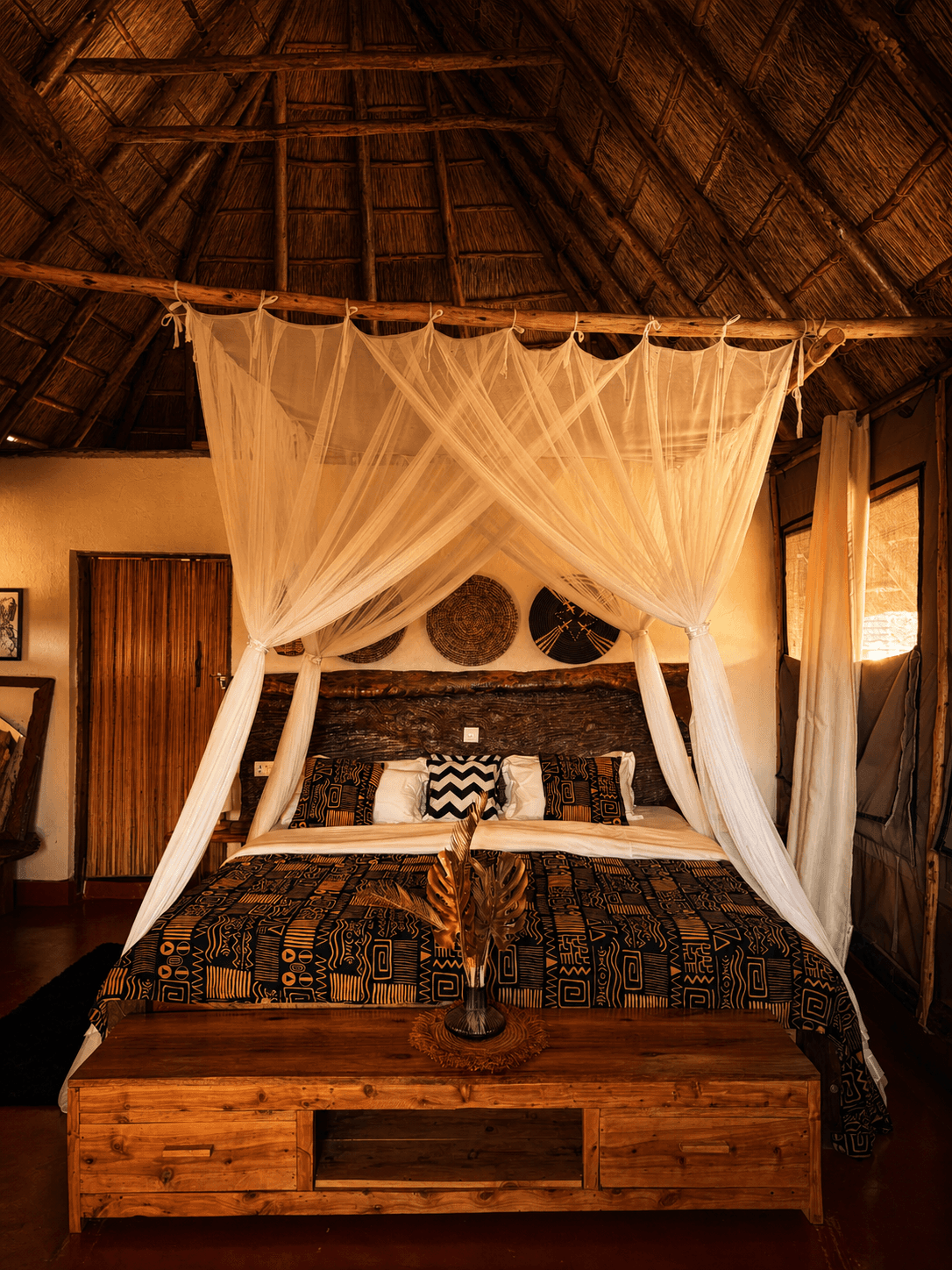 Luxury Safari Tent bedroom at The Elephant Oasis — four-poster canopy bed with white mosquito net, driftwood headboard, African mudcloth bedding, woven basket wall art, thatched ceiling, Murchison Falls Uganda